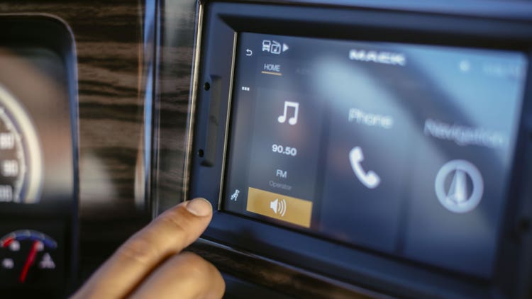 Close-up of a Mack truck's infotainment touchscreen displaying music, phone, and navigation options, with a hand interacting.