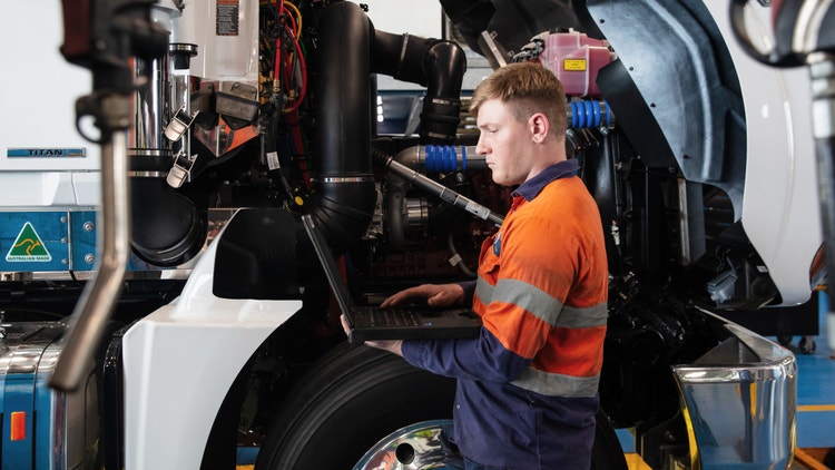 A technician in a high-visibility uniform using a laptop to inspect a Mack truck in a service bay, highlighting protection plans for maintenance and repairs