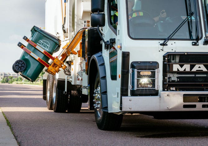 Front view of a Mack LR garbage truck with an automated arm lifting a green waste bin