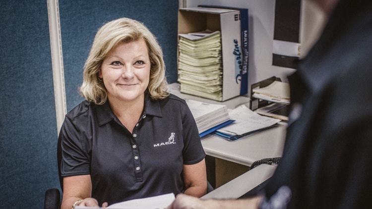 A Mack Trucks employee in a branded polo shirt assisting a customer, with warranty documents on the desk and organized files in the background