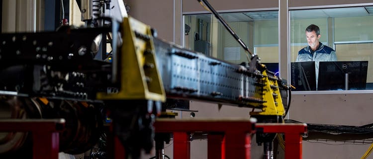 A man working at a computer in a control room, overlooking heavy-duty equipment being tested on a platform inside a workshop